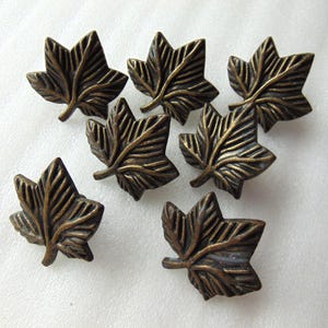 May include: Seven bronze-colored leaf-shaped decorative buttons. Each button has detailed veining and a three-dimensional design. The buttons are arranged on a white surface, showcasing their intricate details and metallic finish.