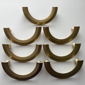 May include: Seven brass cabinet pulls in a semi-circular design. The pulls have a smooth, curved shape with a flat back and rounded ends. The brass has a warm, golden hue, and the pulls are arranged on a white background.
