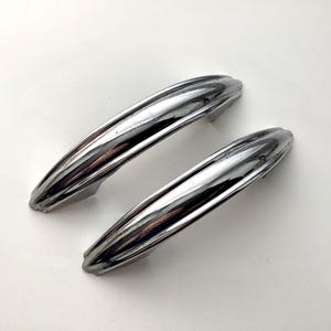 May include: Two vintage chrome door handles with a streamlined, ribbed design. The handles are highly reflective, showing a bright, shiny surface. The handles are likely for a car or cabinet.