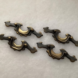 May include: Four antique-style drawer pulls with a dark bronze finish and gold accents. The decorative hardware features a curved design with ornate detailing, suitable for cabinets or furniture restoration projects.