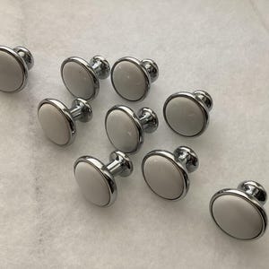 May include: A collection of eight round cabinet knobs featuring a smooth white circular face set within a polished chrome metal frame. Each knob has a short cylindrical base and a rounded top, displaying a consistent design across all pieces.
