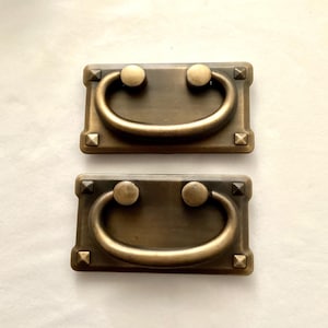 May include: Two antique bronze drawer pulls. Each pull has a rectangular backplate with a curved handle and two round knobs. The backplates have decorative pyramid-shaped studs at each corner. Hardware for furniture restoration.