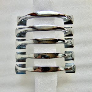 May include: Five polished chrome cabinet handles. Each handle has a curved, ergonomic design with a central twist. The handles are arranged vertically against a white background, showcasing their reflective surfaces.