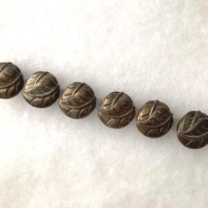 May include: A row of six round metal beads, each featuring a raised leaf pattern with a dark, antiqued bronze finish. The beads are strung together on a thin, light-colored cord, displaying a consistent circular shape and detailed texture.