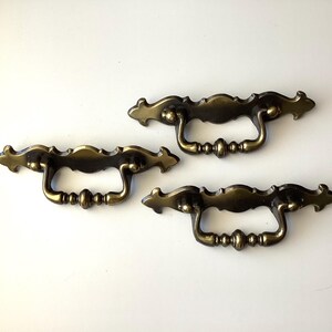 May include: Three antique brass drawer pulls with a decorative, ornate design. Each pull features a curved handle and detailed embellishments. The pulls are a warm, aged brass color, perfect for vintage furniture restoration.