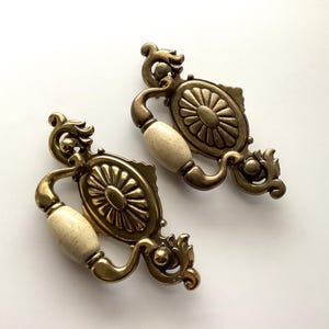 May include: Two antique brass cabinet pulls with cream-colored ceramic handles. The pulls have an ornate design with a central oval medallion featuring a sunburst pattern. The brass has a warm, aged finish, and the ceramic handles have a speckled appearance.