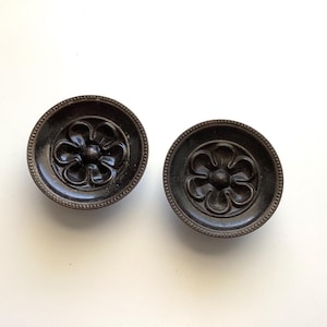 May include: Two dark brown, circular cabinet knobs with a floral design in the center. The knobs have a beaded edge and are set against a white background. These decorative knobs are likely for furniture.