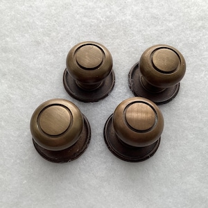 May include: A set of four round cabinet knobs with a brushed antique brass finish. Each knob features a smooth, circular center disc surrounded by a raised ring, mounted on a flat, circular base plate that provides a stable attachment point.
