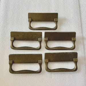 May include: Five antique brass drawer pulls with a rectangular shape and a curved handle. The pulls have a worn, aged finish.
