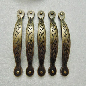May include: Five antique bronze cabinet pulls with a leaf design. Each pull has a curved, elongated shape with a decorative leaf pattern etched down the center. The pulls are approximately 5 inches long and have a circular end on each side.