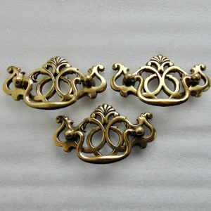 May include: Three antique brass drawer pulls with ornate, symmetrical designs. Each pull features a curved handle with decorative flourishes and a central, stylized floral element. The hardware is in a warm, aged brass finish.