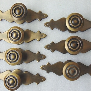 May include: Six antique brass cabinet knobs with ornate, decorative plates. Each knob has a round, raised center with a concentric circle design.