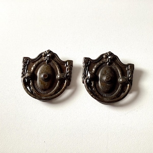May include: Two antique bronze-toned drawer pulls. Each pull has a decorative shield shape with an oval center and ornate detailing. The pulls are likely made of metal and are designed for furniture restoration or decorative use.