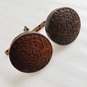 May include: Two antique bronze cufflinks with intricate, raised relief designs. The circular faces feature detailed patterns, and the connecting bars show signs of age. The cufflinks are set against a white background.