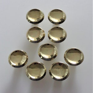 May include: Ten polished brass cabinet knobs arranged on a white surface. Each knob has a rounded, flat top and a slightly curved base. The knobs are evenly spaced, reflecting light.
