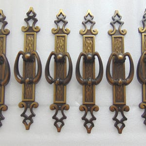 May include: Six antique brass cabinet door pulls with ornate designs. Each pull has a rectangular plate with a decorative pattern and a ring-shaped handle.
