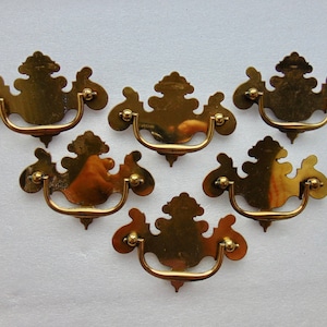 May include: Six ornate brass drawer pulls with a decorative, floral design. Each pull has a curved handle and a decorative backplate.