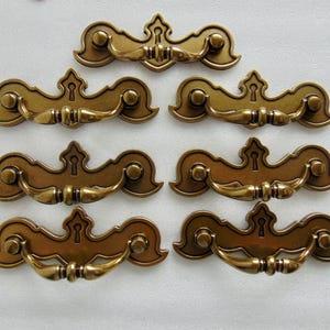 May include: A collection of seven antique-style drawer pulls. Each pull is made of bronze-colored metal with a decorative design. The pulls feature a keyhole detail and curved handles, suitable for furniture restoration or new builds.