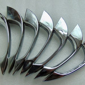 May include: A set of six chrome-plated door handles. Each handle has a curved design with a pointed end. The handles are arranged in a row, reflecting light and creating a shiny appearance. The handles are likely for a car or other vehicle.