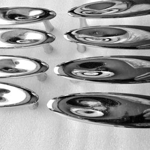 May include: A collection of eight polished chrome cabinet pulls. Each pull has an oval handle with a curved design, mounted on two small, cylindrical posts. The pulls are arranged in two rows of four, reflecting light.