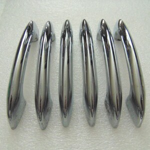 May include: A set of six identical chrome-finished metal drawer pulls arranged in a vertical row. Each handle features a smooth, curved, and elongated design with a reflective surface, tapering slightly at both ends for a sleek appearance.