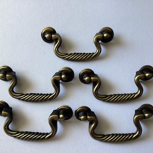 May include: Six antique brass drawer pulls with a twisted rope design on the handle. The pulls have a curved shape and are mounted on a white surface. These decorative pulls are ideal for furniture restoration.