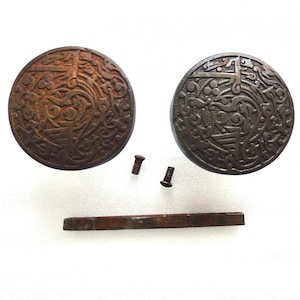 May include: Two circular metal door knobs featuring an intricate, raised relief pattern of swirling lines and geometric shapes. One knob has a rusted, reddish-brown finish, while the other is dark grey. Includes two small metal screws and a flat metal bar.