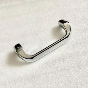 May include: A shiny, chrome-finished cabinet handle with a curved, rectangular design. The handle is a single piece, with a smooth, reflective surface. The ends are slightly thicker than the center. The handle is designed for easy gripping.