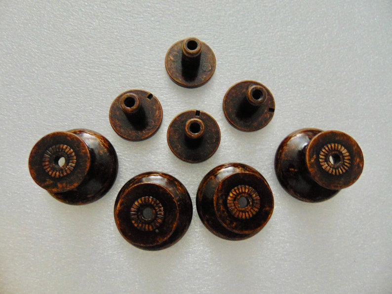 May include: Eight antique bronze cabinet knobs with a decorative design. The knobs have a round base and a raised center with a small hole in the center.