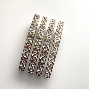 May include: Four bronze-colored cabinet pulls with a decorative scroll design. The pulls are rectangular with a white background and a detailed pattern. The hardware is ideal for adding a vintage touch to furniture.