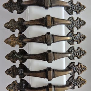 May include: Six antique bronze-toned drawer pulls. Each pull features a decorative, ornate design on the ends and a central bar for gripping. The pulls are arranged vertically against a white background, showcasing their aged finish and intricate details.