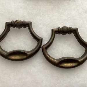 May include: Two matching antique brass drawer pulls with a curved, fan-like shape. Each handle features a central spherical detail on the top bar and a smooth, rounded grip area at the bottom, showcasing a weathered, dark metallic patina finish.