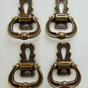 May include: Four antique bronze-colored drawer pulls. Each pull features a decorative backplate with a keyhole design and a curved handle. The pulls have an ornate, vintage aesthetic, suitable for furniture restoration or adding a classic touch.
