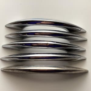May include: A stack of five polished chrome cabinet pulls. Each pull has a curved, elongated shape with a reflective surface. The pulls are arranged horizontally, with a white surface visible between them. The overall design is sleek and modern.