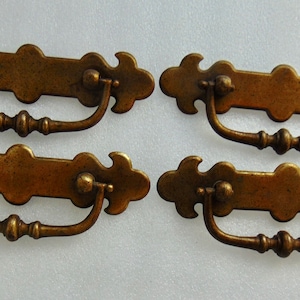 May include: Four antique bronze drawer pulls. Each pull has a rectangular backplate with decorative edges and a curved handle. The pulls are made of metal and have a vintage, aged appearance. They are likely for furniture restoration or a similar project.