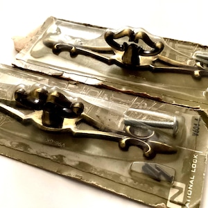 May include: Two vintage brass-toned cabinet pulls in their original packaging. Each pull features a decorative handle with a curved design and includes mounting screws. The packaging is a clear plastic blister pack on a cardboard backing.