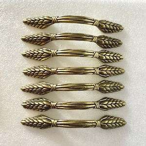 May include: Seven antique brass cabinet pulls. Each pull features a wheat stalk design on the ends and a straight bar in the middle. The pulls are arranged in a neat, parallel row, showcasing their decorative and functional design.