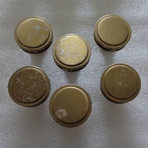 May include: Six vintage brass-colored knobs with a circular design. The knobs have a slightly worn appearance, with some areas showing signs of wear and tear. The knobs are arranged in a circular pattern.