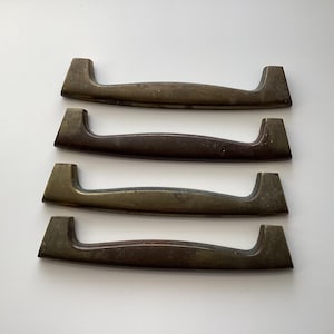 May include: Four vintage bronze-toned cabinet handles. Each handle has a curved bar with rectangular end pieces. The handles are arranged horizontally against a white background, showcasing their aged patina and design.