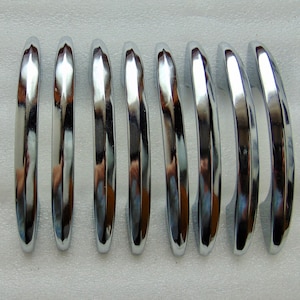 May include: Eight chrome-finished, curved metal pieces arranged in a row. Each piece has a smooth, reflective surface, catching the light and creating highlights. The pieces are evenly spaced and have a rounded, elongated shape.