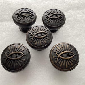 May include: Five bronze-colored cabinet knobs. Each knob features a sunburst design with a fish symbol in the center. The knobs have a rounded shape and a dark, antique finish, suitable for furniture or cabinetry.