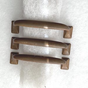 May include: Three matching cabinet pulls made of a smooth, brushed bronze-toned metal. Each handle features a slightly arched, rounded grip section that connects to two square-shaped mounting bases on either end, creating a uniform hardware set.