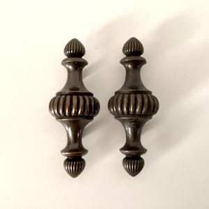 May include: Two antique bronze-colored decorative finials. Each finial has a bulbous, ribbed center section, a slender neck, and a pointed top and bottom. The finials are symmetrical and appear to be made of metal.