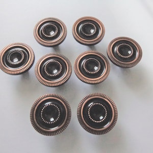 May include: Eight bronze-colored cabinet knobs with a dark center and a decorative, ridged outer ring. The knobs are arranged on a light gray surface, showcasing their circular design and metallic finish.