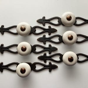May include: Six decorative cabinet pulls. Each pull features a white ceramic knob with a copper-colored center, mounted on a dark bronze-colored metal backplate. The backplates have an ornate, symmetrical design, adding a vintage touch to the hardware.