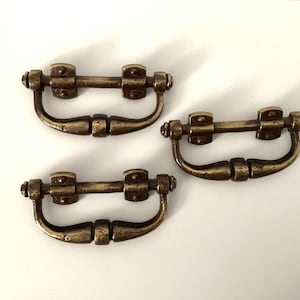 May include: Three antique bronze-colored drawer pulls. Each pull has a curved handle and a rectangular mounting plate with two screw holes. The hardware has a distressed finish, suggesting age or vintage style.