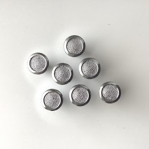 May include: Seven silver-toned cabinet knobs with a textured, dotted center design. The knobs are round with a raised edge and are arranged on a white surface. These decorative knobs are suitable for furniture or home decor projects.