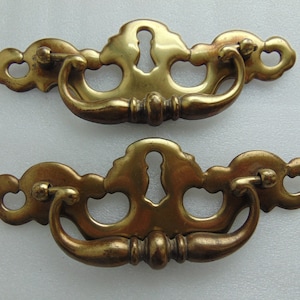 May include: Two antique brass drawer pulls. Each pull features a keyhole design and a curved handle. The pulls have an aged, tarnished appearance, suggesting they are vintage or vintage-inspired. The hardware is likely for cabinets or furniture.