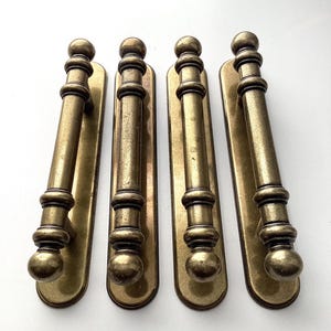 May include: Four antique brass door pulls. Each pull features a long, cylindrical handle with decorative ball and ring accents, mounted on a rectangular backplate. The brass has a warm, aged patina, suggesting vintage or antique hardware.