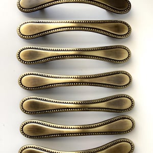 May include: Seven antique brass cabinet pulls. Each pull has a curved, elongated shape with a beaded edge and a slightly darker finish on the ends. The pulls are arranged in a vertical row against a white background.